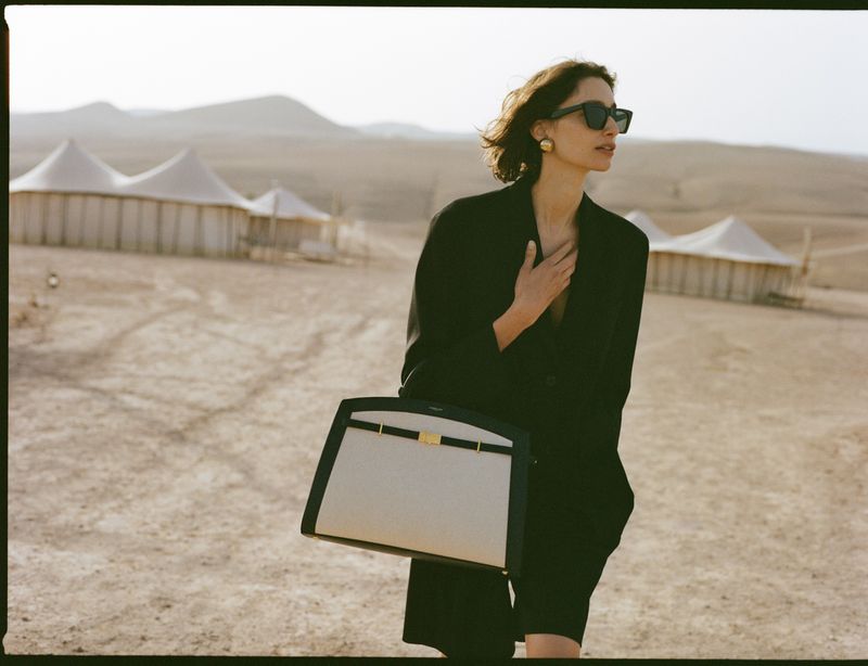 Model in black jacket carrying a two-tone handbag walking across a sandy desert landscape