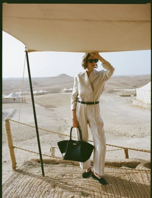 Model in cream suit holding a black leather tote bag under a desert canopy for DeMellier London S/S 2025 campaign by Sonia Szóstak.