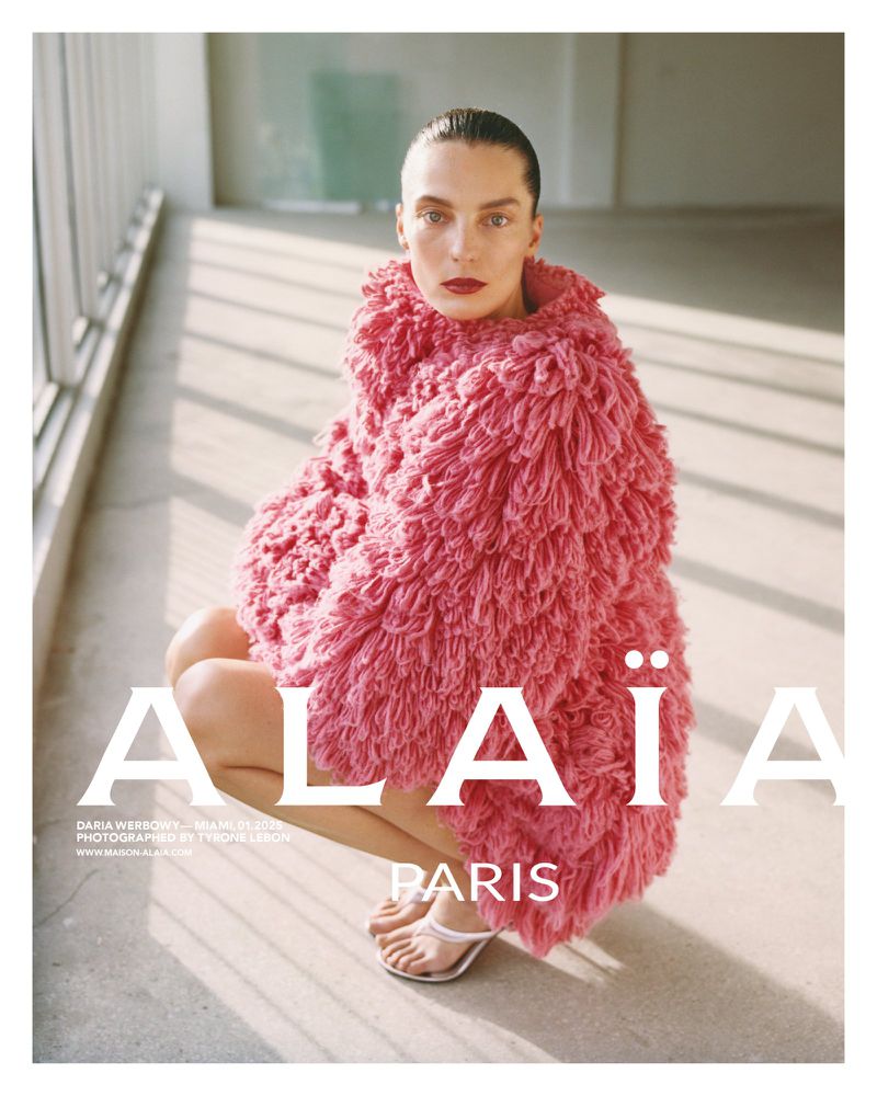 Fashion image of a woman squatting, wearing a pink faux fur coat. The background is a simple gray concrete wall and floor.