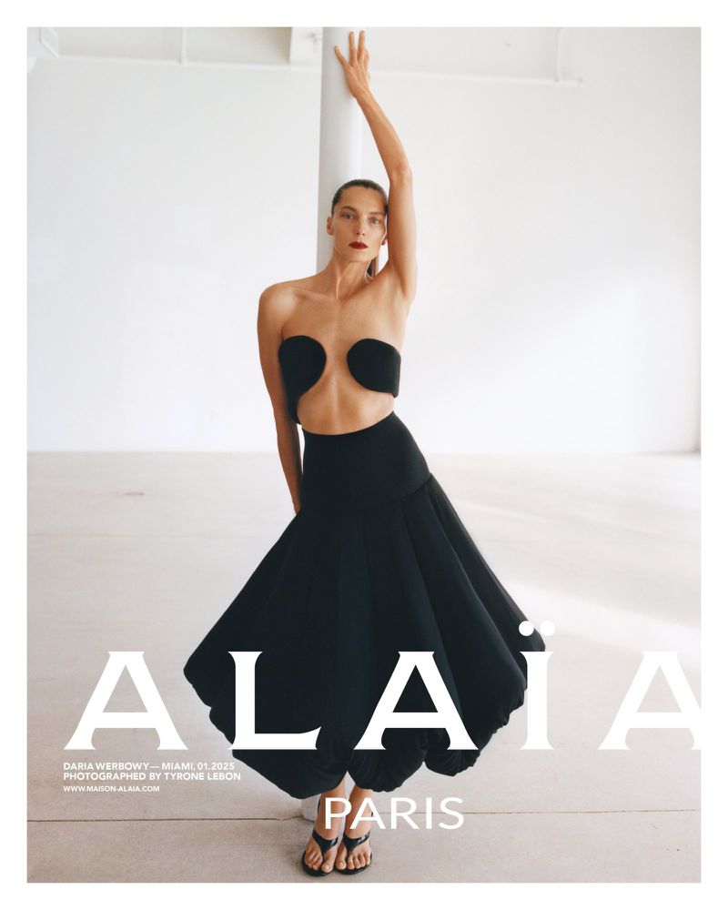 Fashion editorial shot of a female model in a black strapless top and full skirt, posing in a minimalist photo studio setting.
