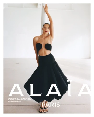 Fashion editorial shot of a female model in a black strapless top and full skirt, posing in a minimalist photo studio setting.