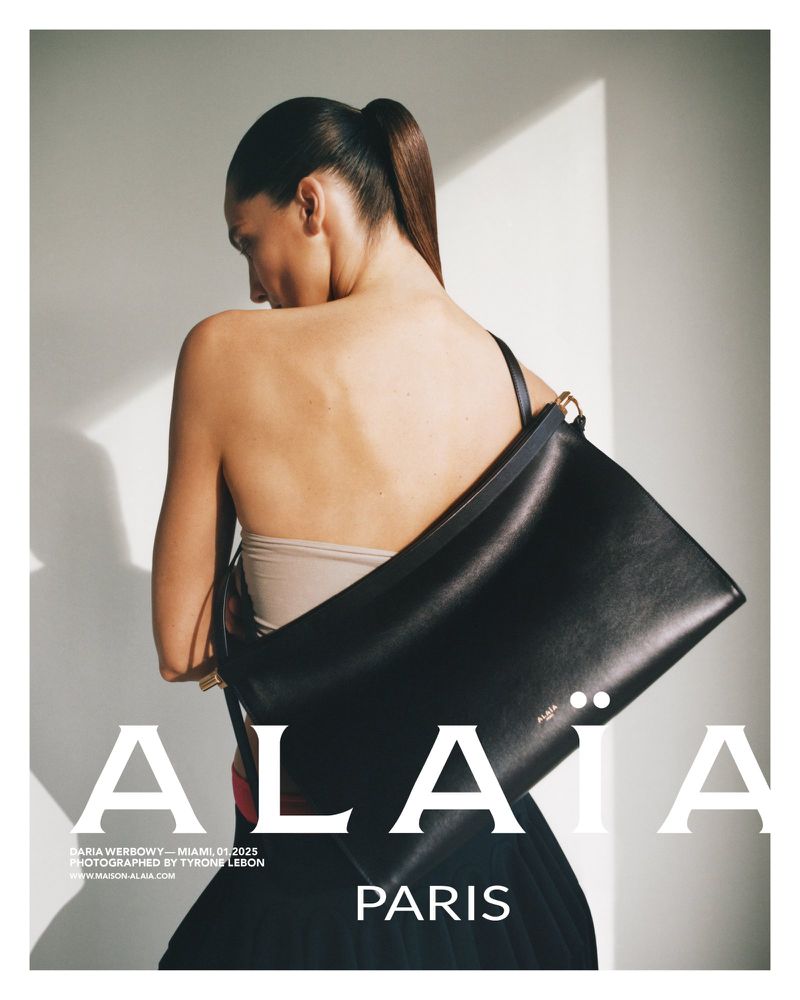 A fashionable adult female poses in a studio with a large black leather handbag slung over her shoulder for a fashion editorial.