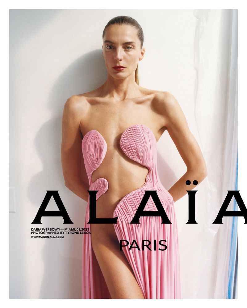 Fashion editorial shot of a woman wearing a pink Alaïa gown posing against a white wall with black Alaïa Paris brand text overlay.
