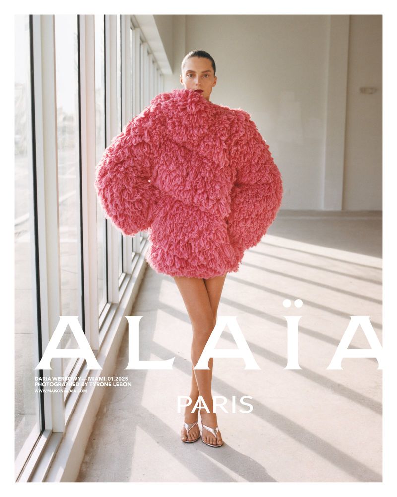 Fashion editorial shot of a female model in a pink, textured coat standing in front of large windows in a well-lit interior space.