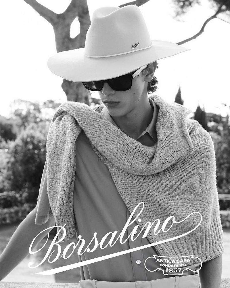 Monochrome shot of a young adult male wearing a hat, sunglasses, and sweater draped over his shoulders, posing in an outdoor setting.