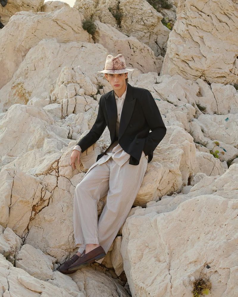 A young adult man wearing a hat, blazer, pants, and shoes is posing on rock formations in outdoor daylight, styled for fashion editorial.