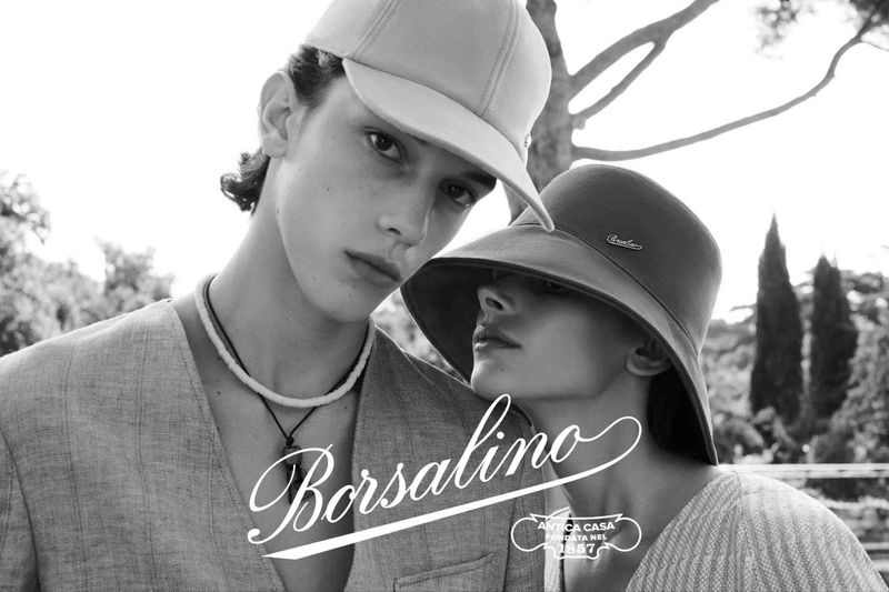 Black and white portrait of a fashionable couple modeling hats, wearing linen clothing in a sun-drenched outdoor setting with greenery.