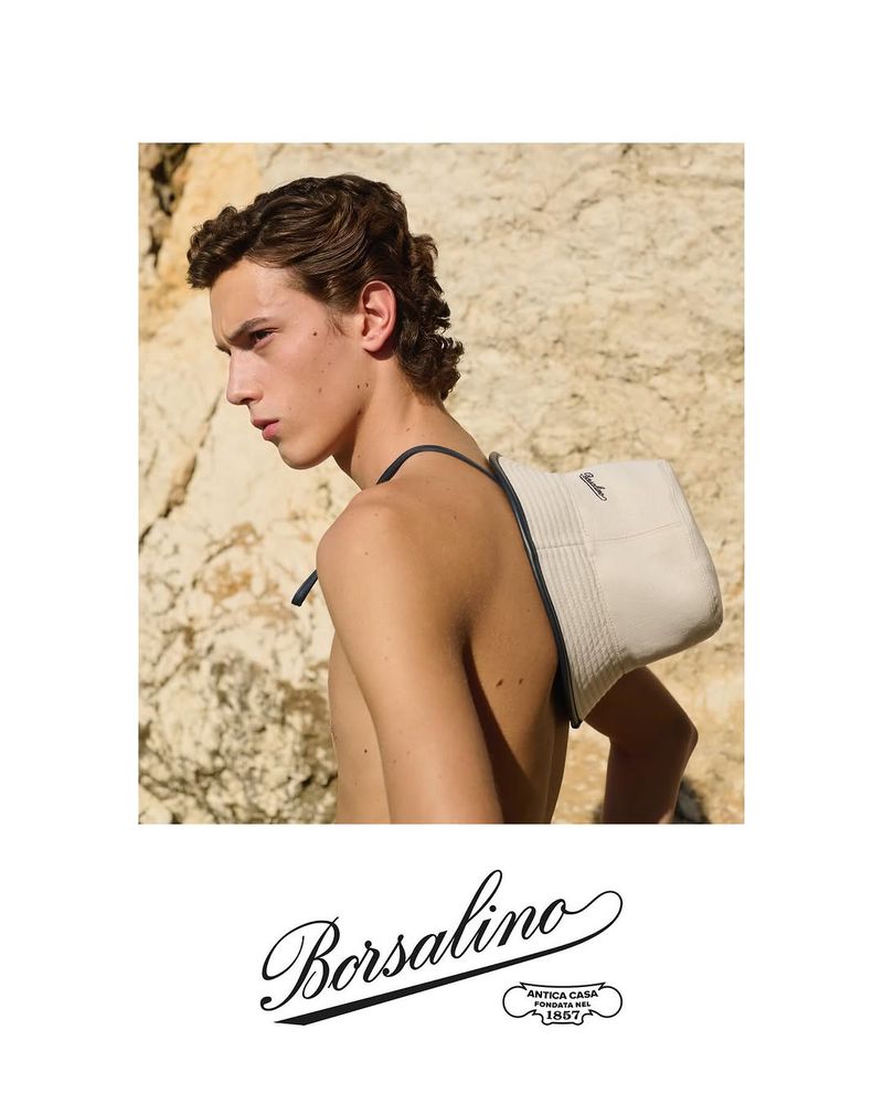 Borsalino brand advertisement featuring a fair-skinned young adult male with a beige bucket hat worn on his back against a rock background.