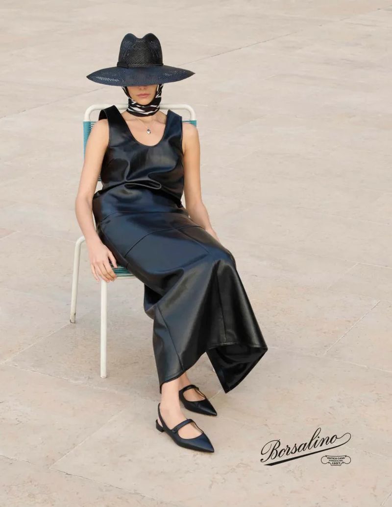 A woman sits outdoors on a chair wearing a black leather dress, a black wide-brimmed hat, and black shoes in a fashion editorial shot.