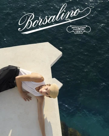 A model poses on a concrete structure overlooking the sea in a Borsalino ad campaign, featuring a tan hat and a white tank top.