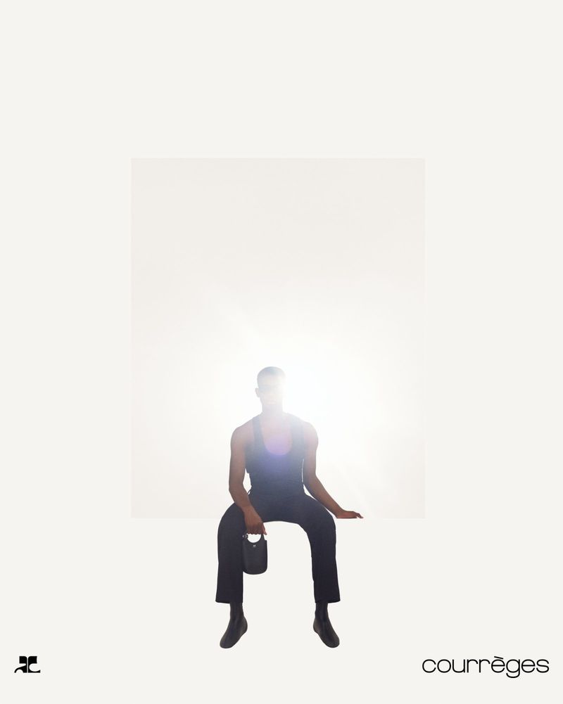 Model seated against a bright white background wearing dark clothing and holding a small handbag