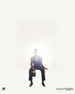 Model seated against a bright white background wearing dark clothing and holding a small handbag