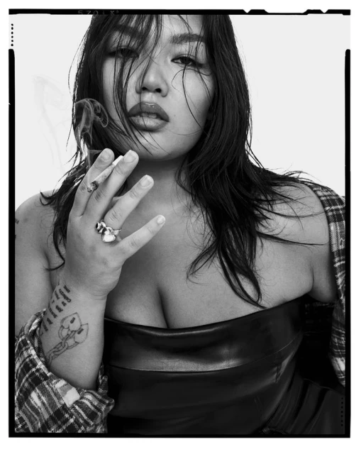 Monochrome portrait of an Asian woman smoking, featuring dark hair, rings, tattoos, leather top, and a checkered shirt over her shoulder.