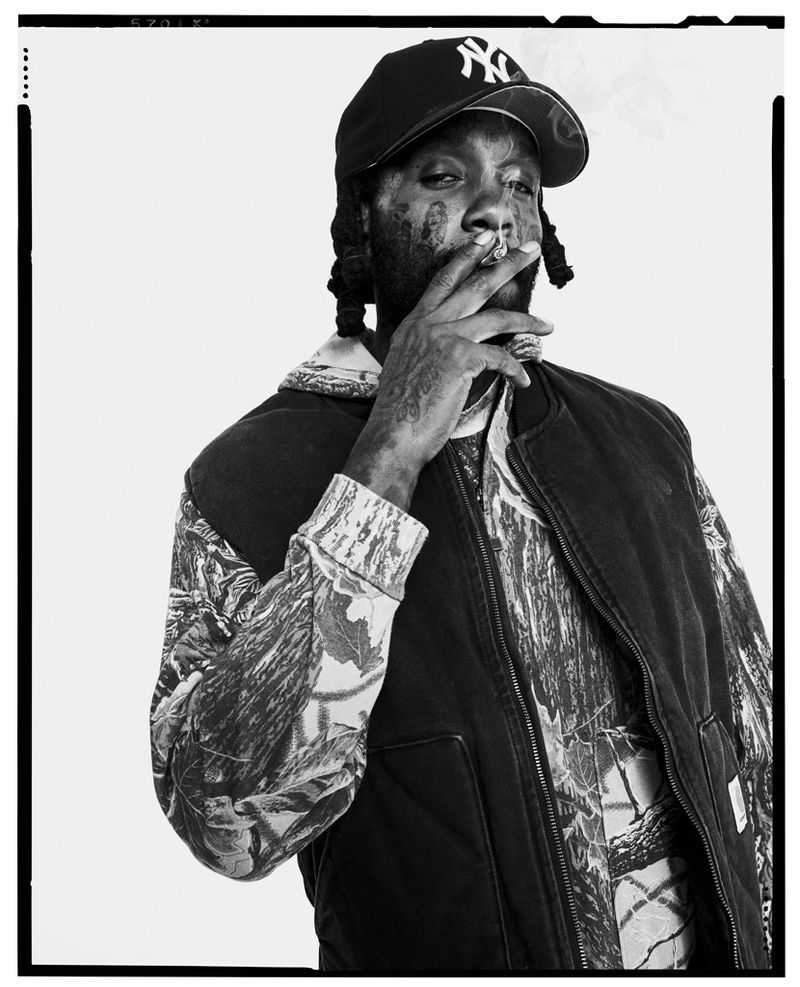 Black and white portrait of a man wearing a baseball cap and patterned jacket, smoking against a white background