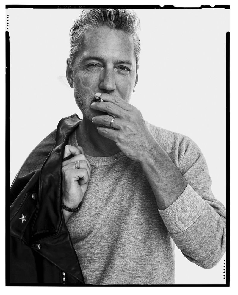 Monochrome portrait of a mature man in a grey sweatshirt, holding a cigarette to his mouth, with a leather jacket over his shoulder.