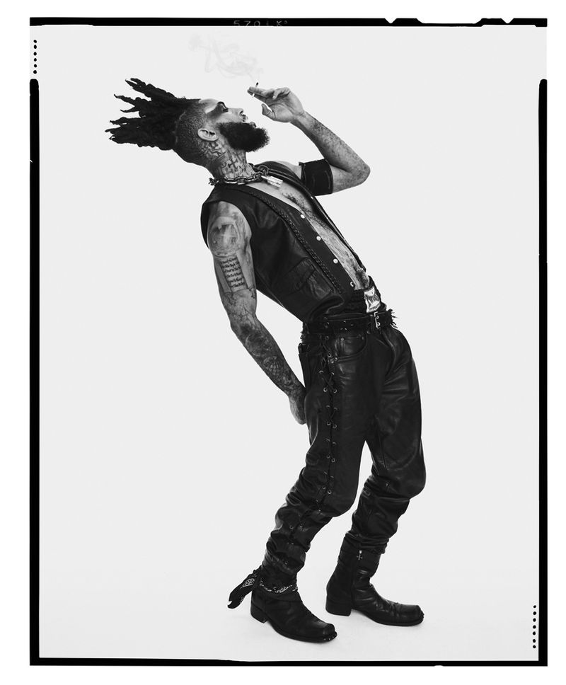 Black-and-white fashion portrait of a tattooed man with dreadlocks wearing leather vest and pants, posing against a white background.