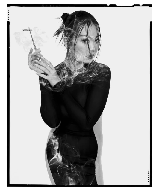 Black and white studio portrait of a woman in a black top holding a long cigarette holder, partially obscured by smoke.