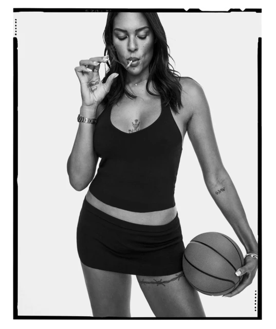 Monochrome studio shot of a woman holding a basketball and holding a cigarette up to her mouth, wearing a cropped tank top and miniskirt.
