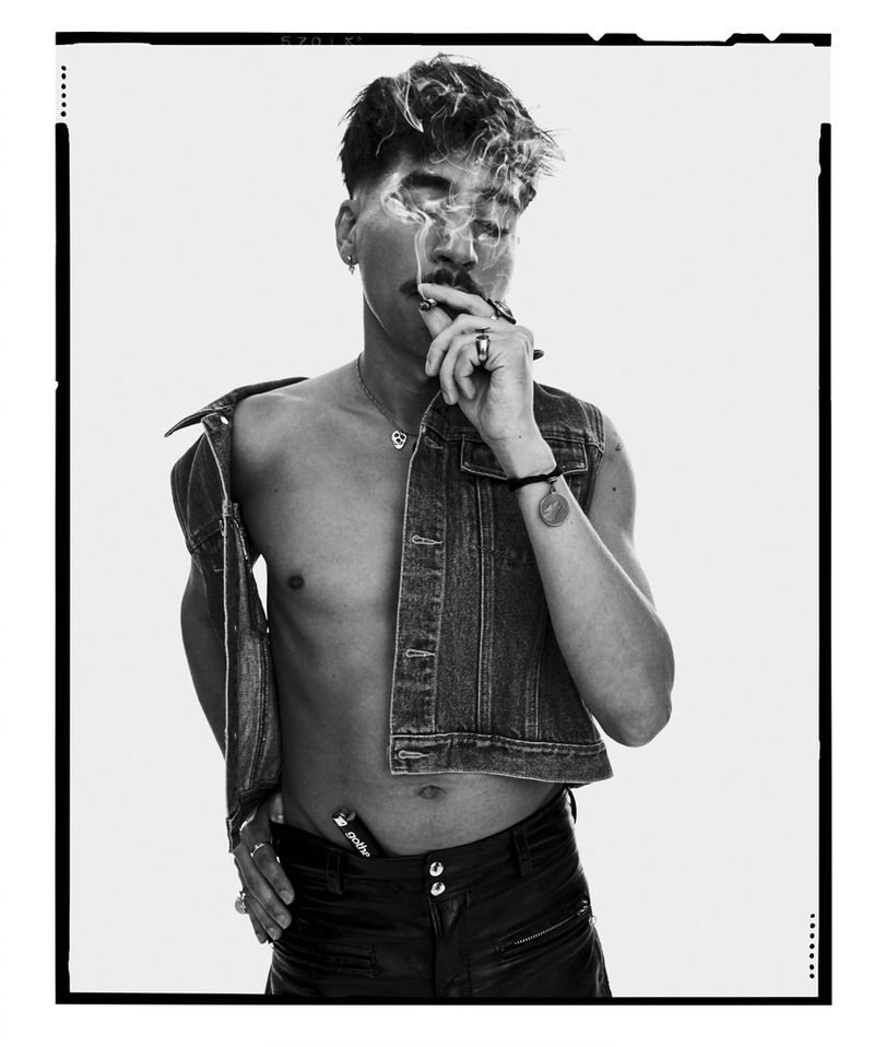 Black and white portrait of a shirtless man with a mustache smoking a cigarette, wearing a denim vest and leather pants against a white backdrop.
