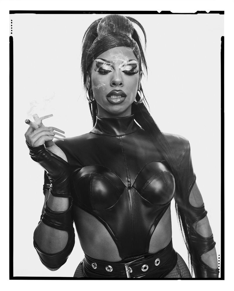 Striking black-and-white studio shot of an adult with dramatic makeup, dark leather outfit, hoop earrings, and smoking a cigarette.