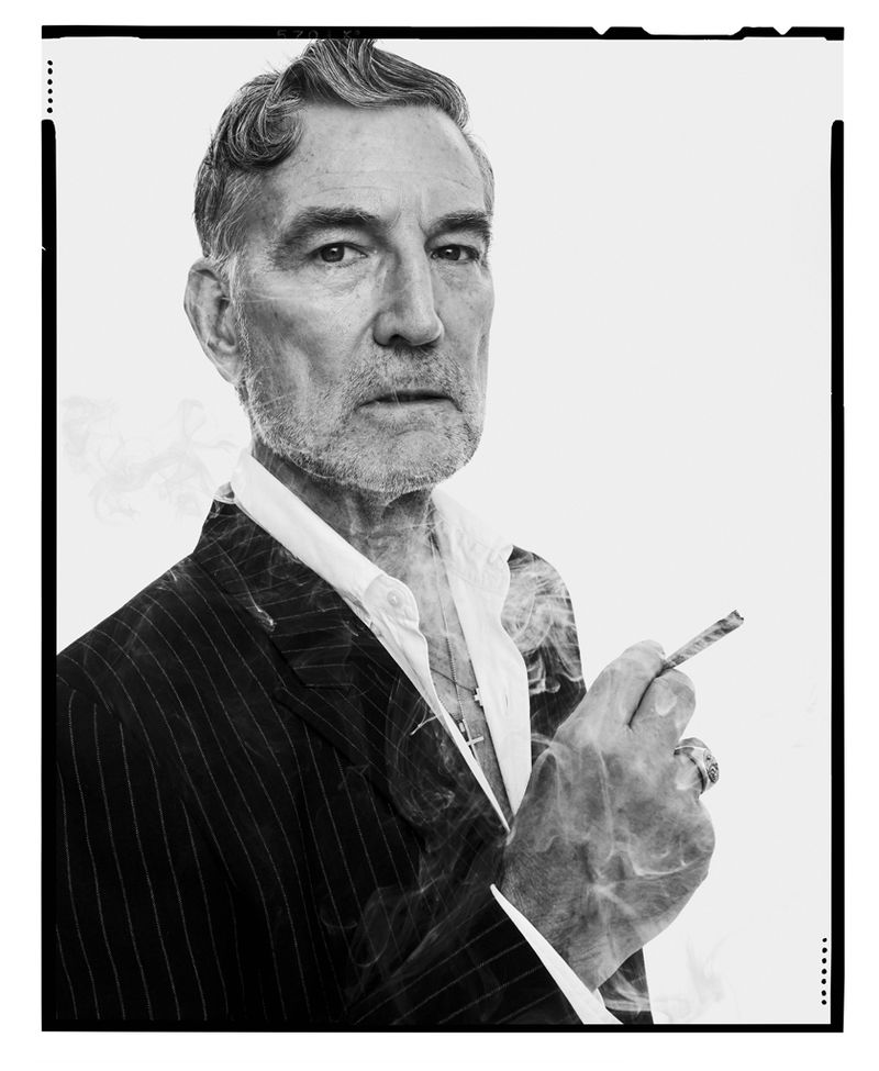 Portrait of an older man with grey hair in a pinstripe suit, smoking a cigarette, against a stark white background. Studio lighting.