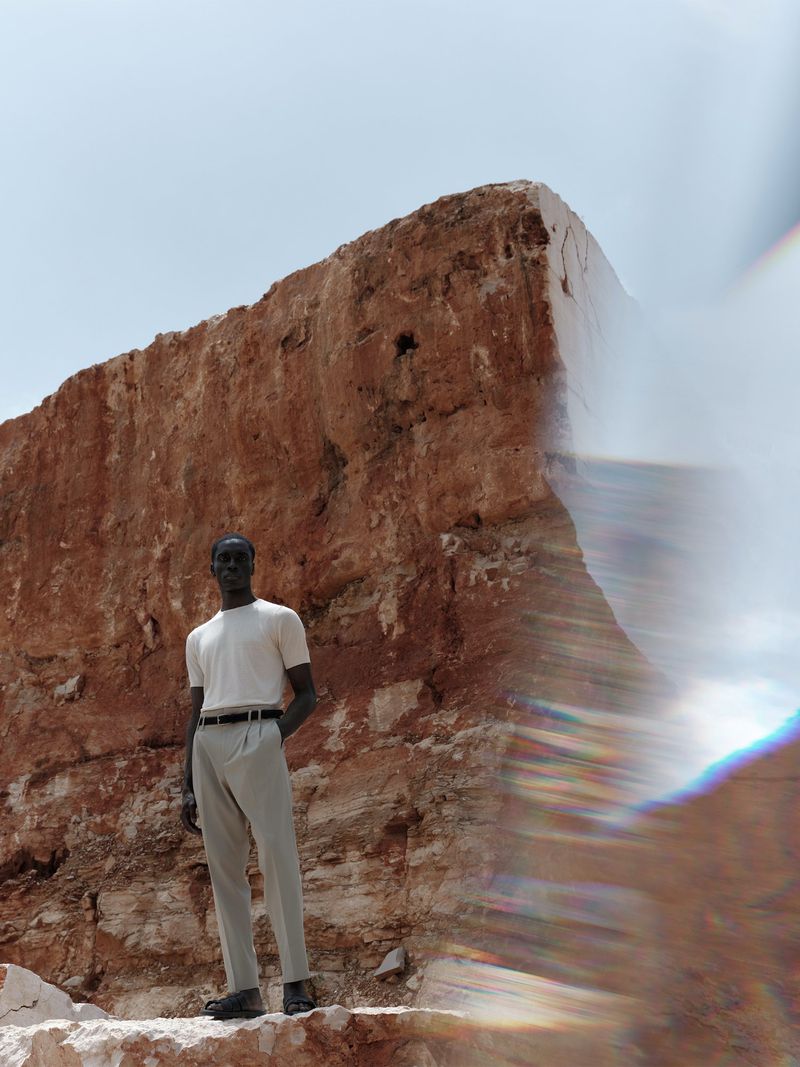 An adult male model in a casual white t-shirt and khaki trousers stands in front of a huge weathered rock formation in daylight.