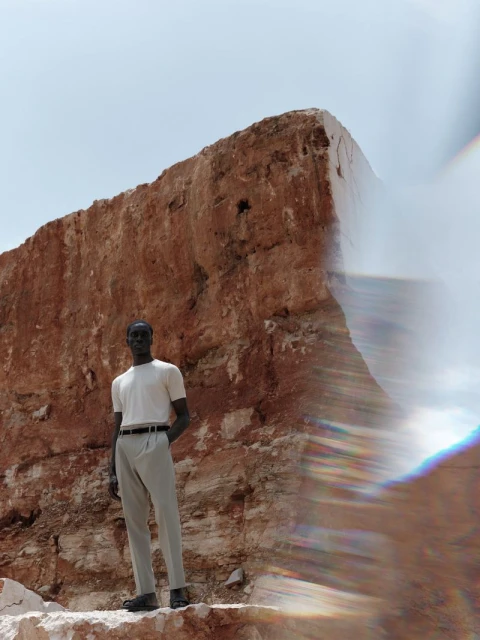 An adult male model in a casual white t-shirt and khaki trousers stands in front of a huge weathered rock formation in daylight.