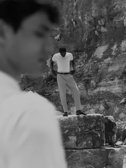 Monochrome outdoor shot with an adult man in light trousers and shirt standing on rock, looking down, a second person in the foreground.