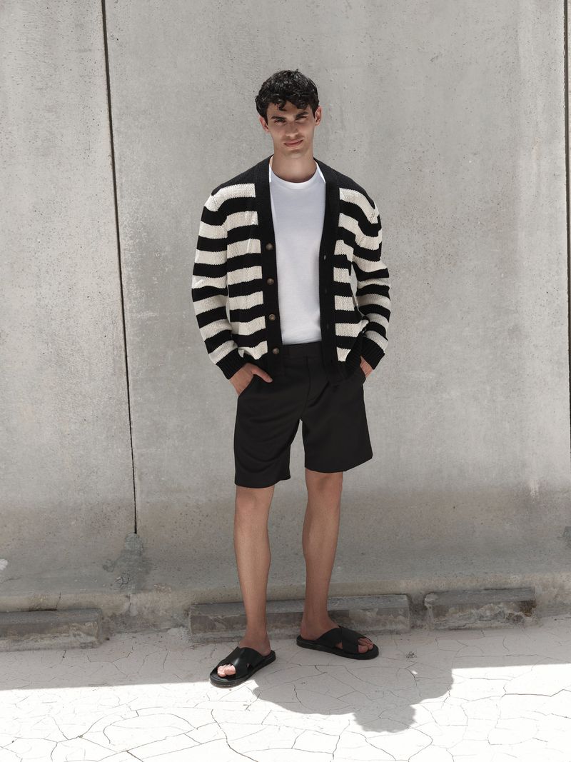Man in striped cardigan, white t-shirt, and black shorts poses against a textured wall, showcasing casual fashion and relaxed style.