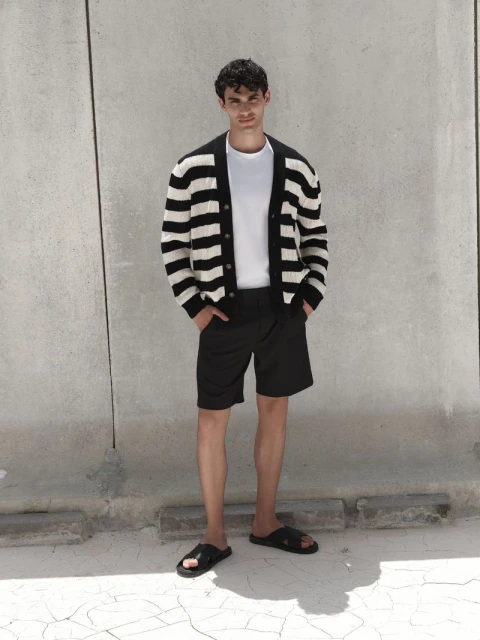 Man in striped cardigan, white t-shirt, and black shorts poses against a textured wall, showcasing casual fashion and relaxed style.