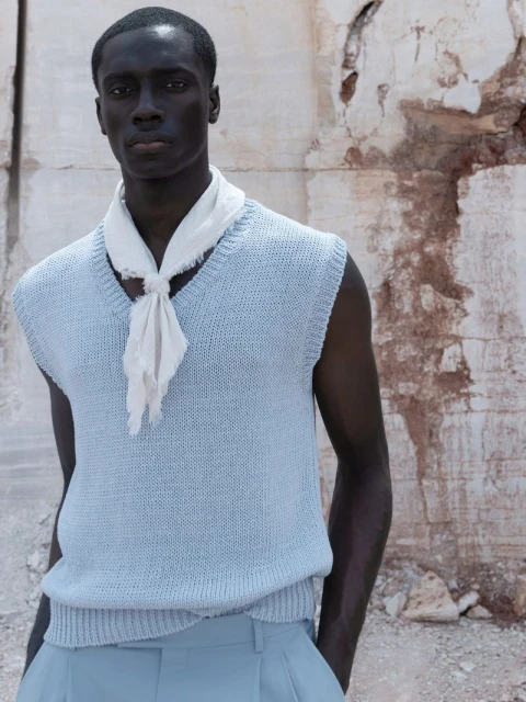 A dark-skinned male model poses outdoors wearing a powder-blue sweater vest and matching pants with a light scarf tied around his neck.