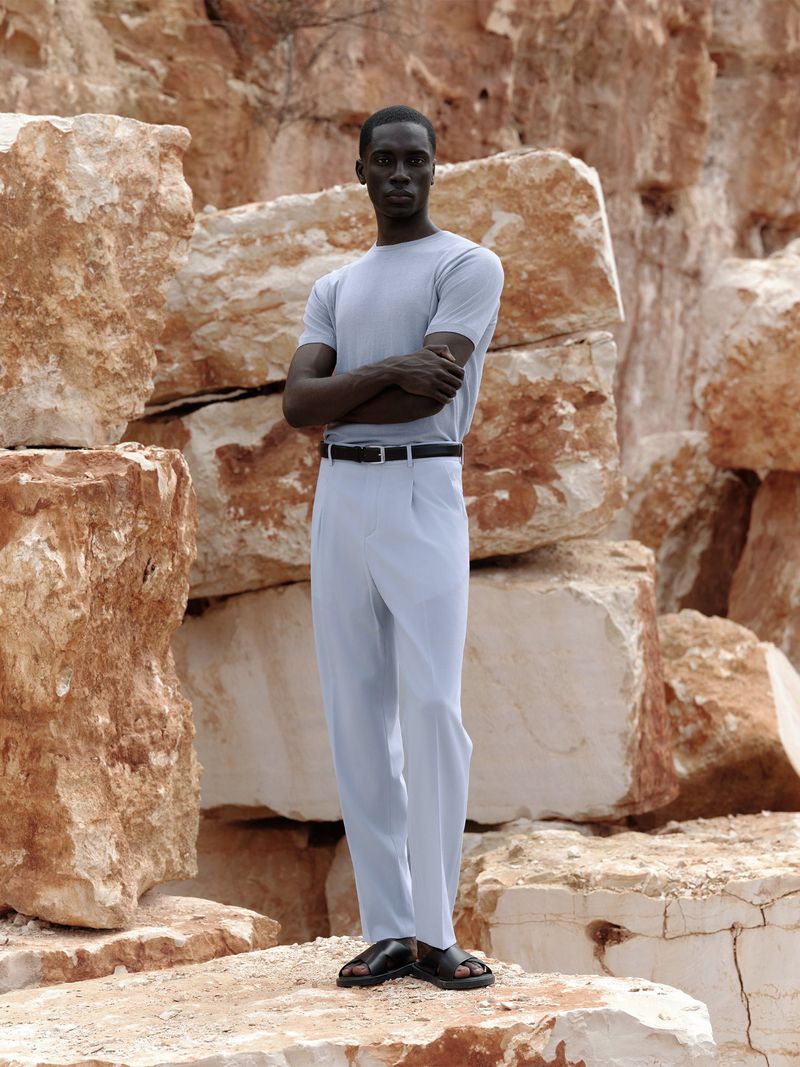 An African-American man stands in light blue shirt and pants, black belt and sandals, posing against a backdrop of large rock formations.