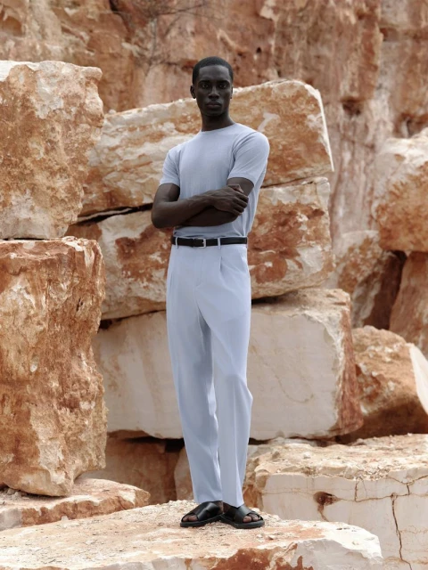 An African-American man stands in light blue shirt and pants, black belt and sandals, posing against a backdrop of large rock formations.