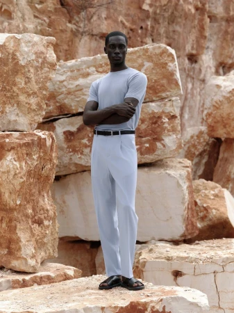 An African-American man stands in light blue shirt and pants, black belt and sandals, posing against a backdrop of large rock formations.