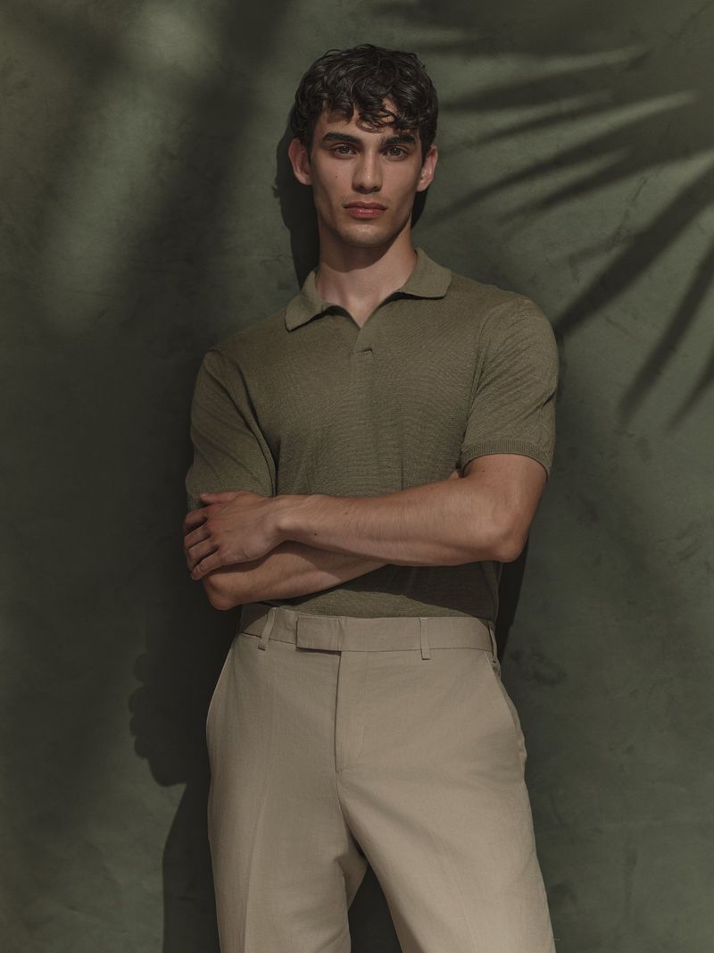 Portrait of a young adult man in a collared shirt and trousers, standing indoors with his arms crossed against a textured wall.