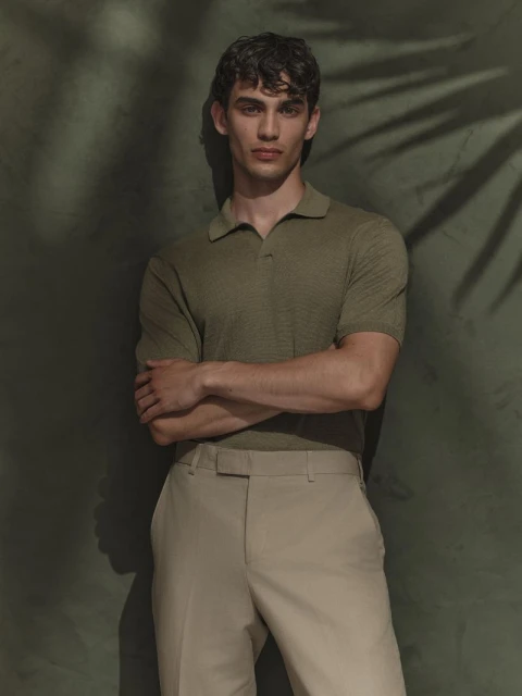 Portrait of a young adult man in a collared shirt and trousers, standing indoors with his arms crossed against a textured wall.