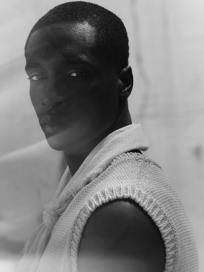 Portrait of an adult black man in a sleeveless knitted top with a scarf, shot in black and white with a shallow depth of field.