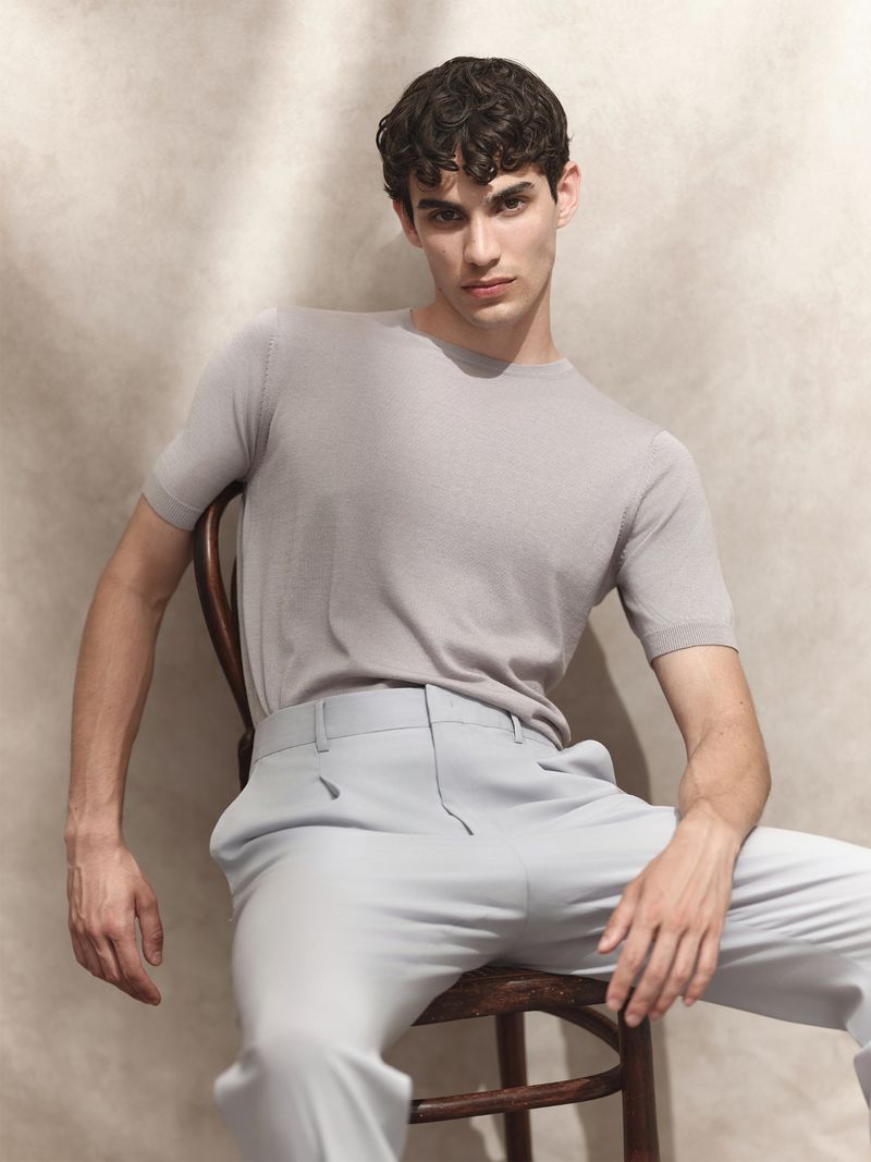 A male model seated on a wooden chair wearing a light grey crewneck top and matching trousers against a textured neutral studio wall.