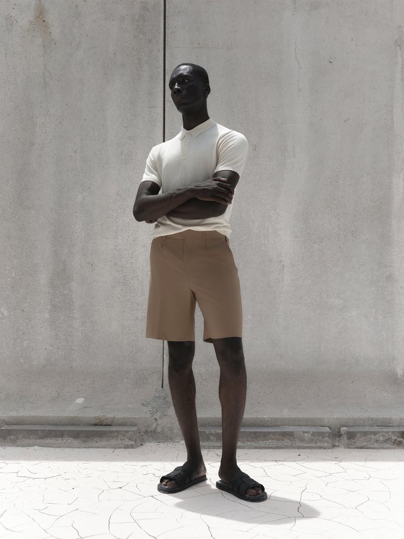 An adult male model with dark skin poses in tan shorts and a white polo shirt against a textured grey wall and cracked white floor.