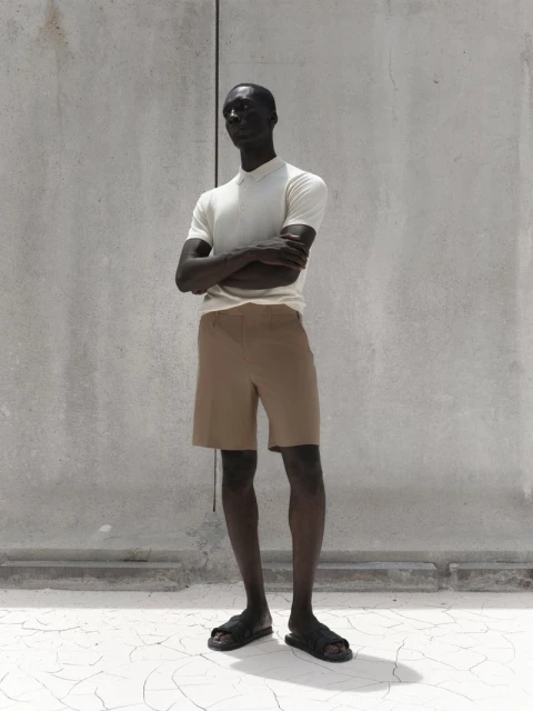 An adult male model with dark skin poses in tan shorts and a white polo shirt against a textured grey wall and cracked white floor.