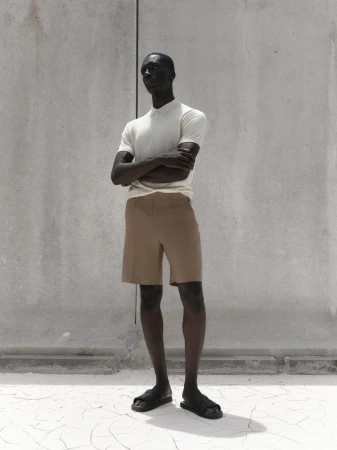 An adult male model with dark skin poses in tan shorts and a white polo shirt against a textured grey wall and cracked white floor.
