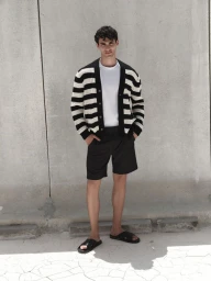 Male model wearing black and white striped cardigan and dark shorts standing against a plain concrete wall.