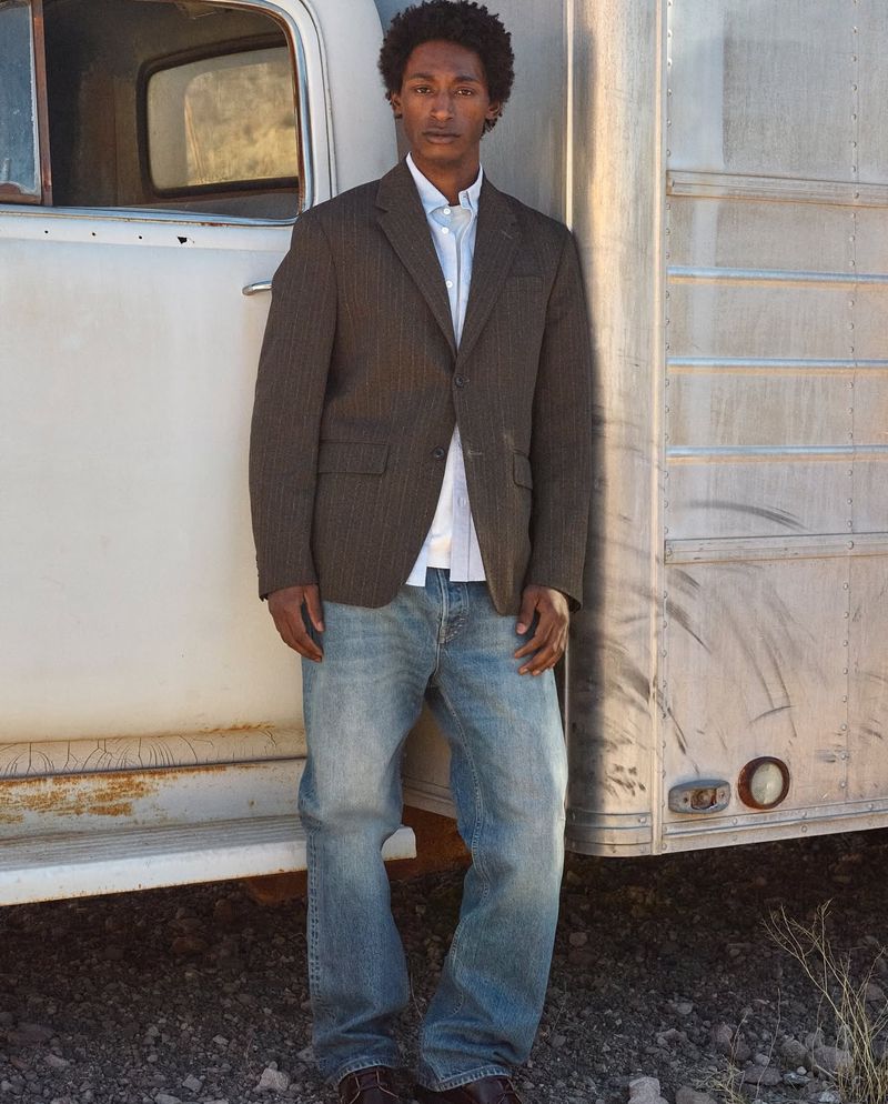 Full shot of a man wearing a brown blazer and jeans, leaning against a silver trailer and looking toward the camera outdoors.