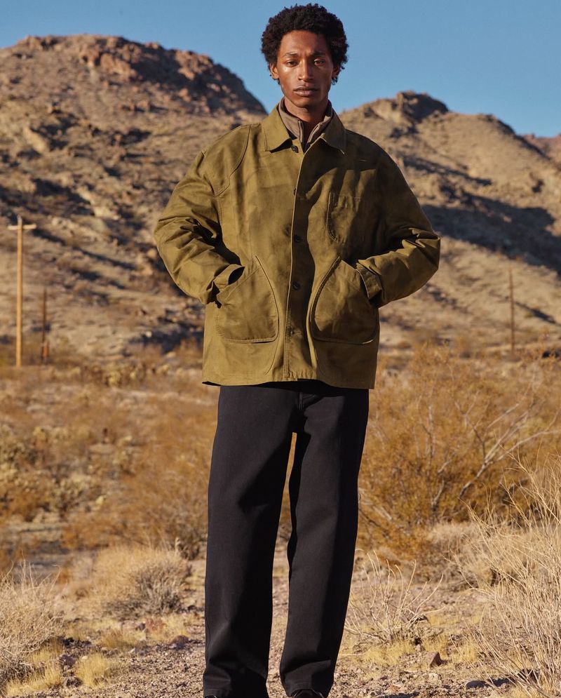 Male model wearing an olive green jacket and black pants stands in a desert landscape with mountains in the background on a sunny day.