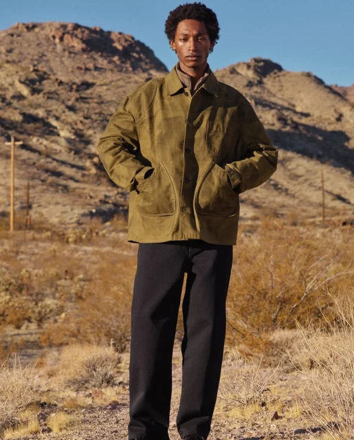 Male model wearing an olive green jacket and black pants stands in a desert landscape with mountains in the background on a sunny day.