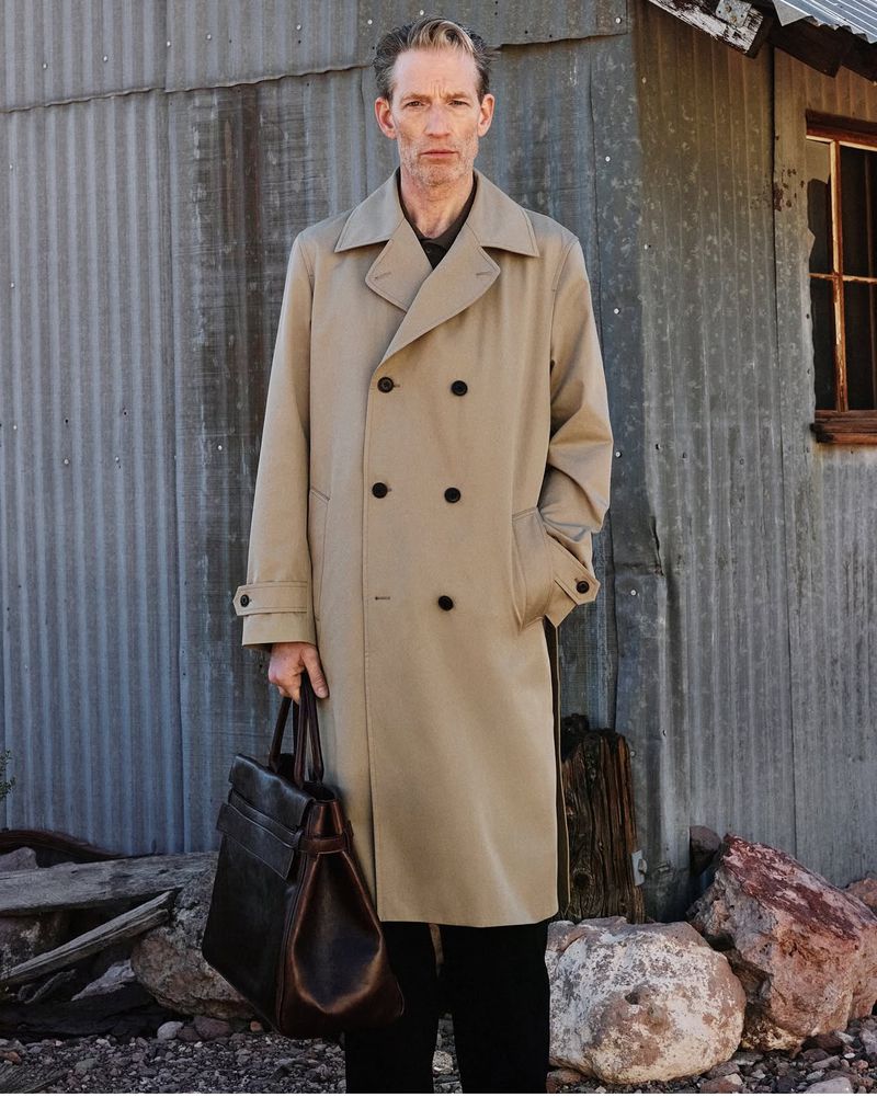An adult male stands in a tan trench coat holding a black leather bag near a weathered corrugated metal building outdoors.