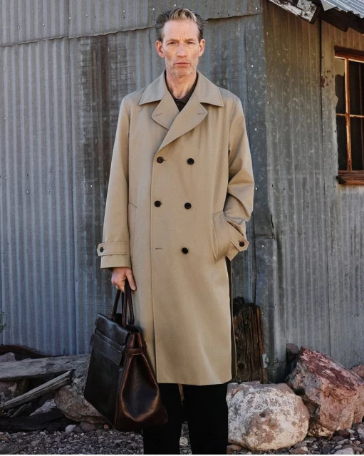 An adult male stands in a tan trench coat holding a black leather bag near a weathered corrugated metal building outdoors.