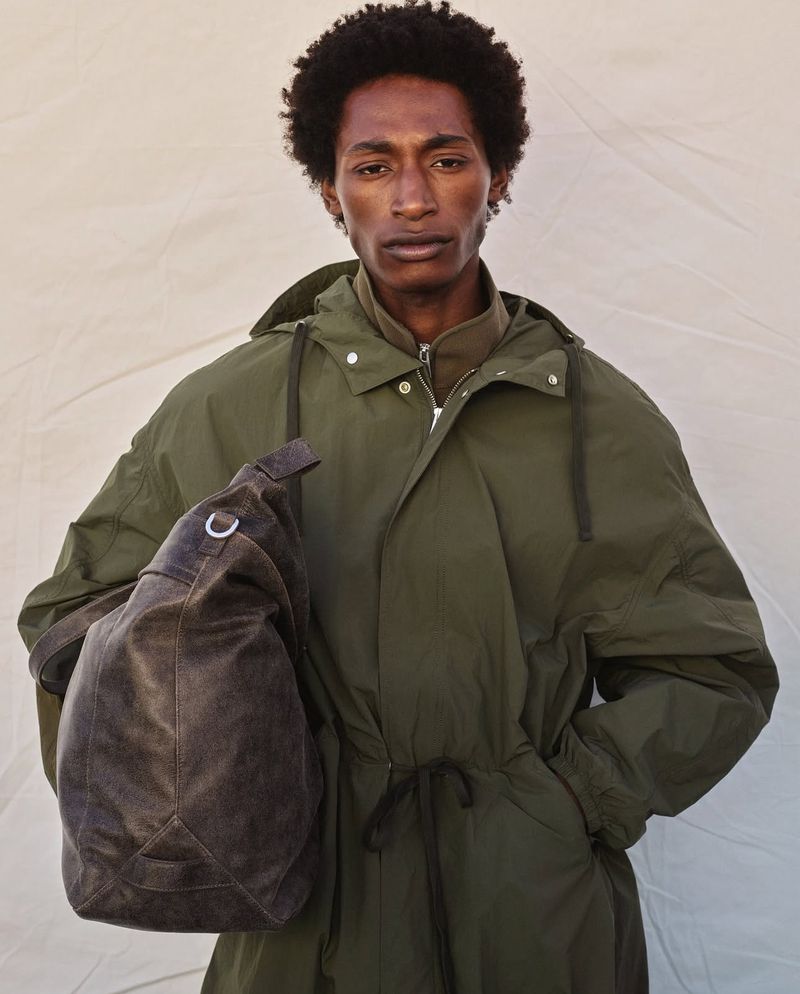 An African-American man poses in an olive green jacket, holding a brown bag on his shoulder in a studio setting with neutral backdrop.