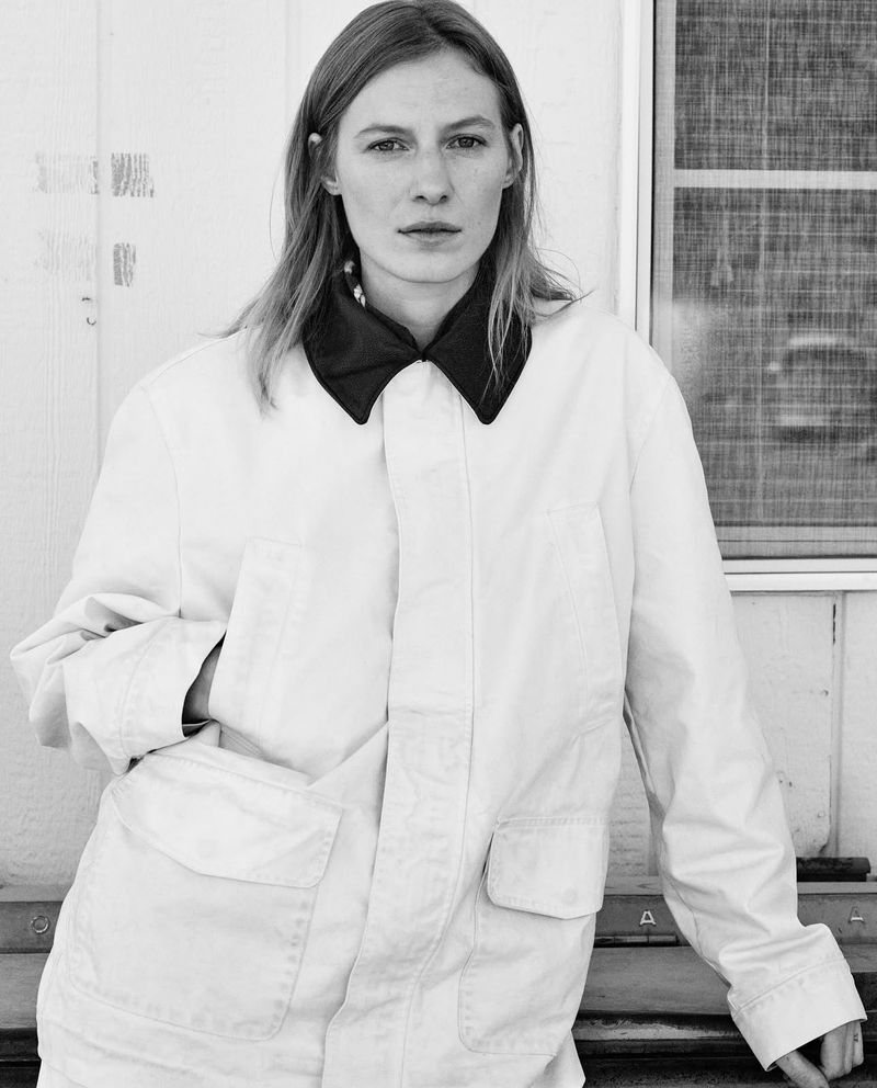 Black and white portrait of a woman wearing a white jacket with a dark collar leaning against a white wall with a screened window.
