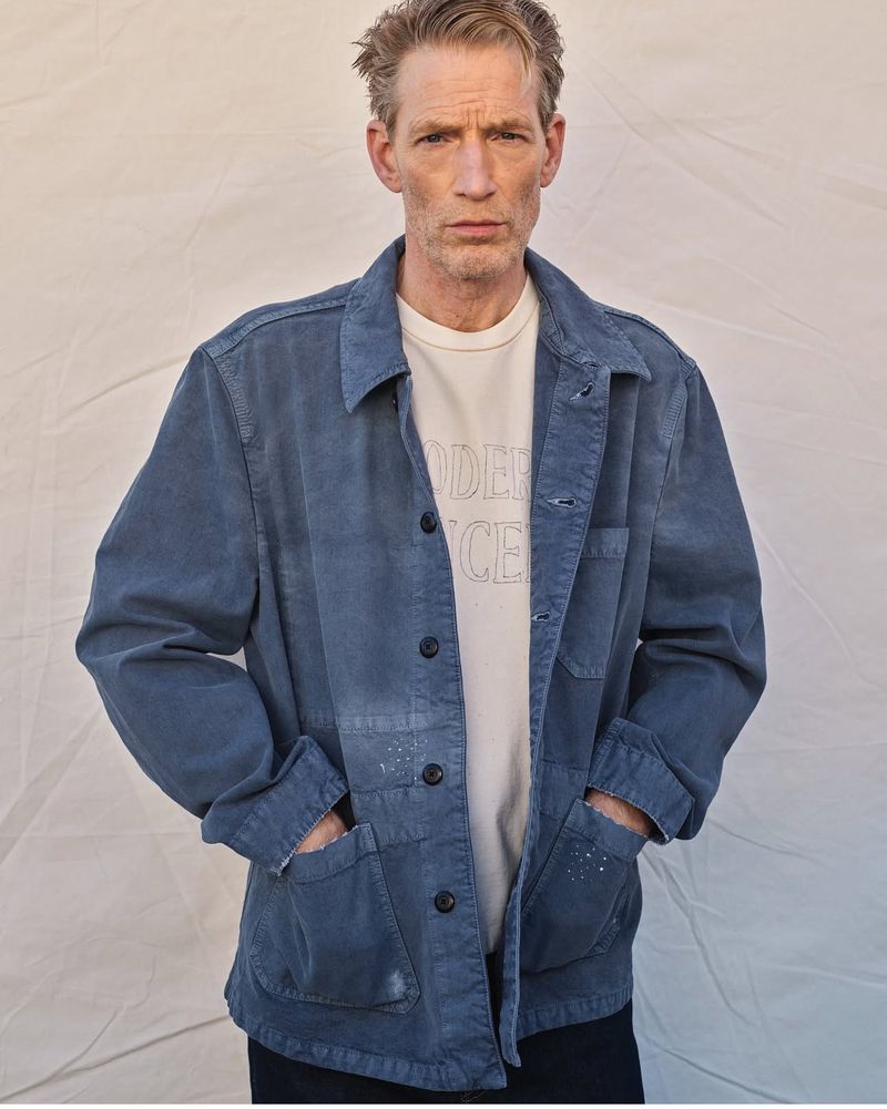 Portrait of a fair-skinned adult male wearing a blue jacket and off-white t-shirt against a neutral, slightly wrinkled background.